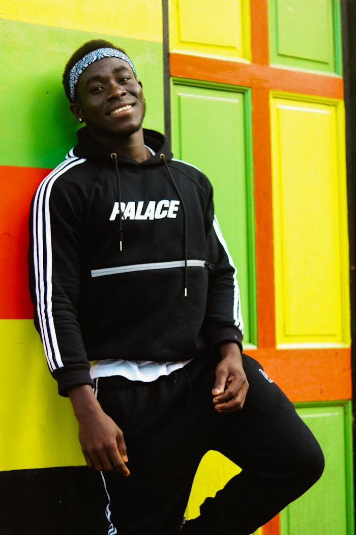 A smiling young man wearing a black hoodie leans against a vibrant, colorful wall outdoors.